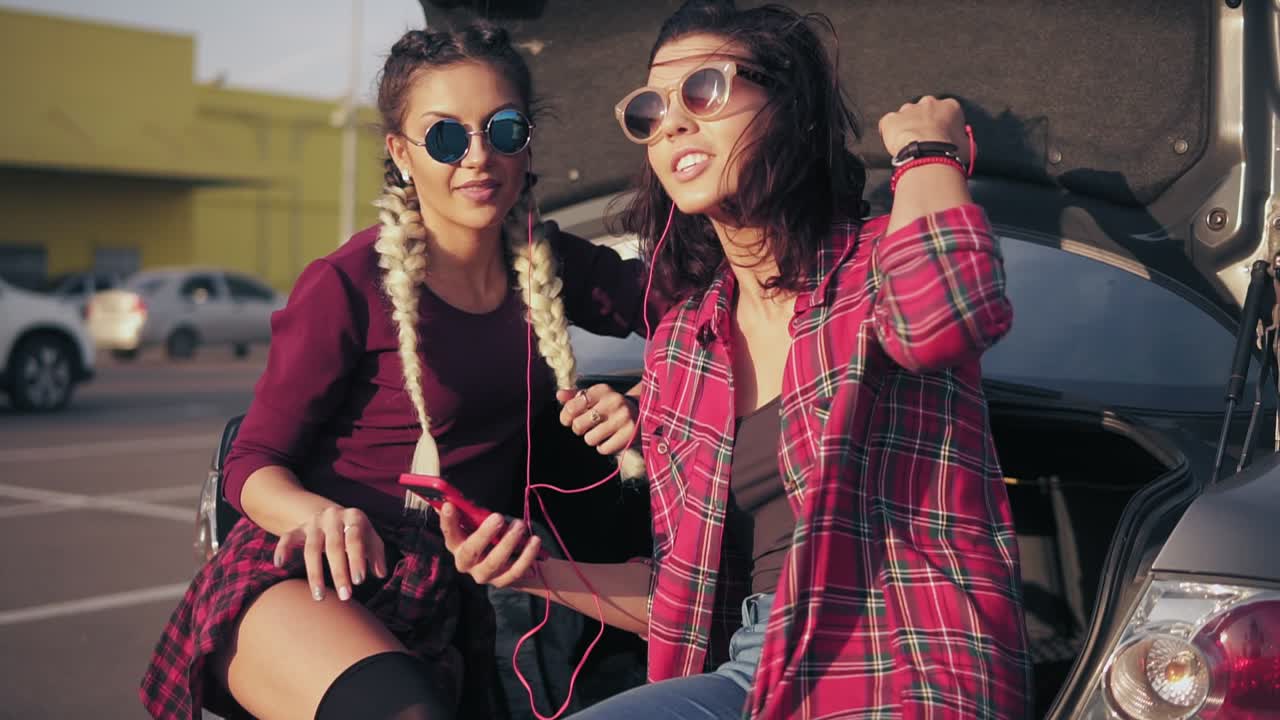 Two young attractive girlfriends in sunglasses sitting inside of the open car trunk in the parking by the shopping mall and