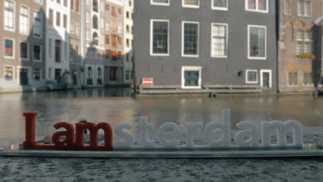 Vista-De-La-Pequeña-Figura-Plástica-De-La-Escultura-De-Letras-De-Iamsterdam-En-El-Puente-Contra-El-Paisaje-Urbano-Borroso-De-Amsterdam,-Países-Bajos