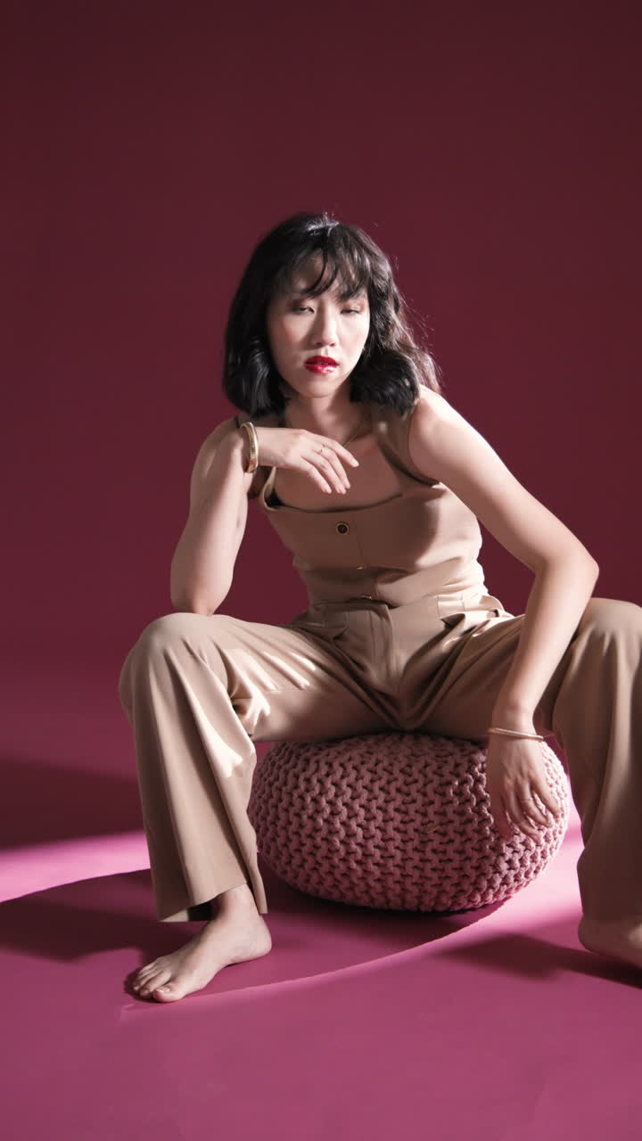 Vertical video: Fashion model in beige outfit sitting on pouf, striking thoughtful pose