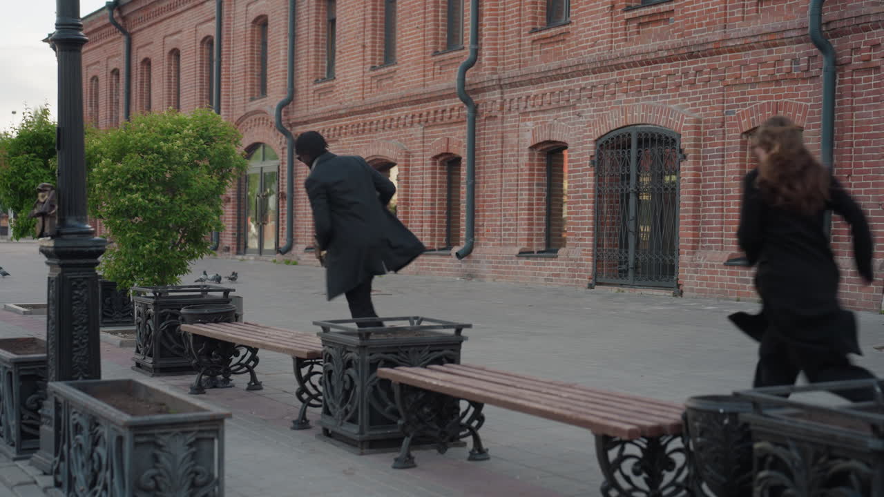 Man in dark coat and eyeglasses interacts with lady sitting on bench near solid brick wall with large pipes outside while evening light highlights texture of urban street and architectural details