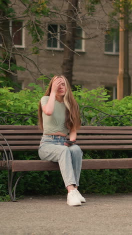 dama sentada en un banco de madera al aire libre, mirando el teléfono en su mano derecha, tocando suavemente su cabello con su mano izquierda, el fondo incluye, árboles y edificio parcialmente oscurecido por el follaje