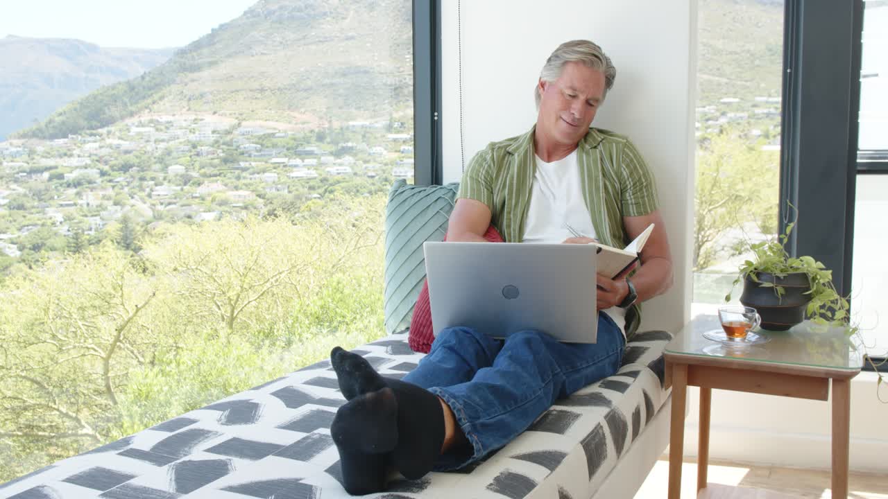 Man reclining on daybed at home with laptop open, noting notebook with pen, working remotely