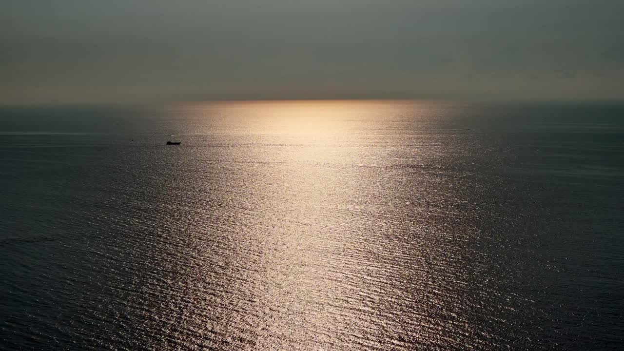 A serene ocean view captured during sunrise, with soft golden light reflecting off the calm water surface. A lone cargo ship is visible in the distance, giving a scale and to the vast seascape