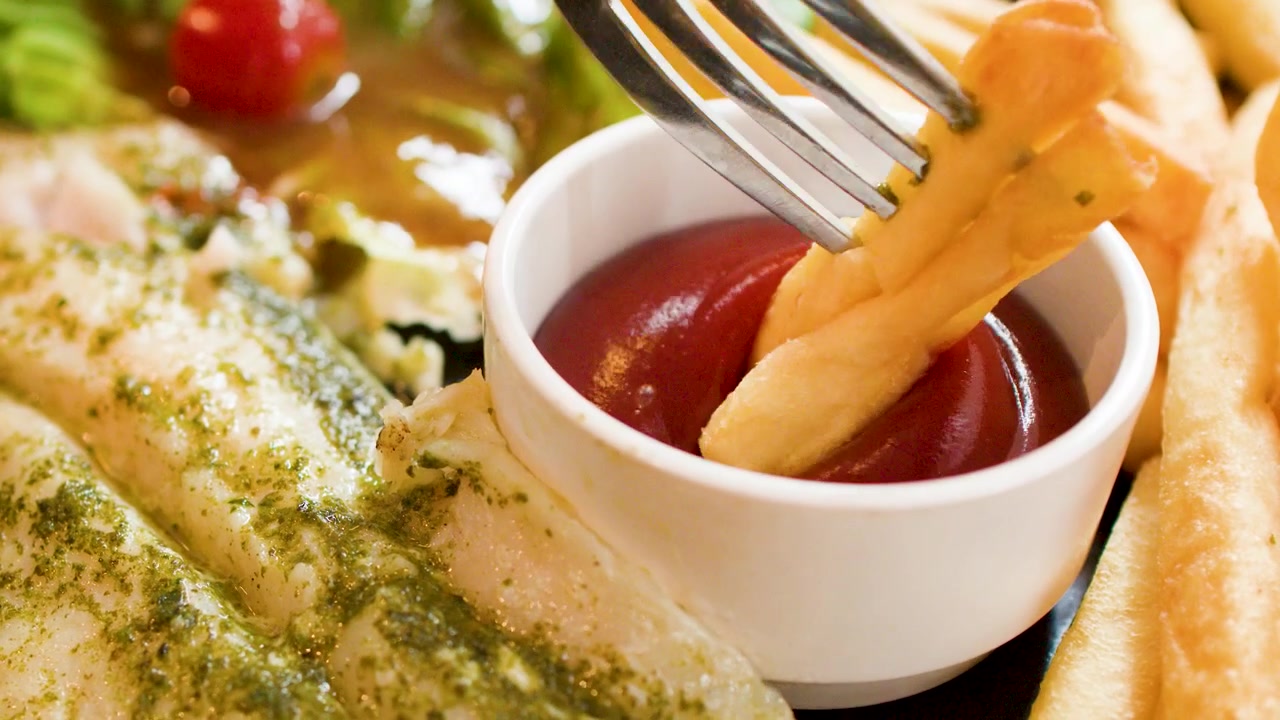 Primo piano di una patatina fritta dorata che si intinge nel ketchup rosso, accanto a pesce alla griglia con pesto.