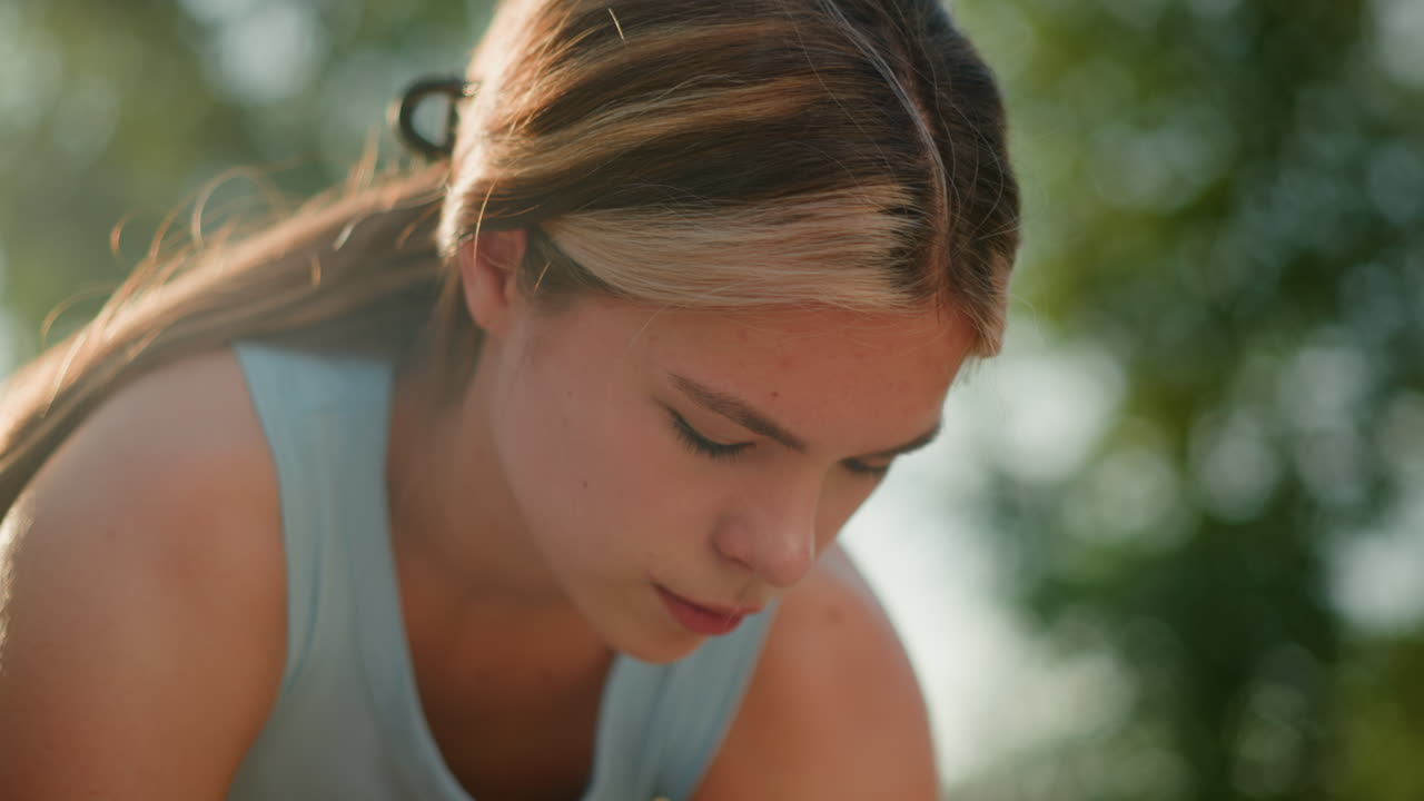 Close-up of young woman slightly bent, focused on something below her, sunlight reflecting off her face, hair tied back, wearing light blue top, with greenery softly blurred in background