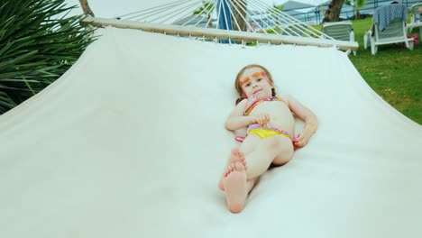 una niña pequeña en traje de baño y con dos coletas tomando el sol en una hamaca de playa y un hotel resort