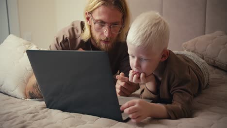 un hombre rubio con gafas y barba, junto con su pequeño hijo albino de ojos azules en una chaqueta marrón, ven una película en una computadora portátil y dibujos animados educativos durante su relajación y pasar tiempo juntos