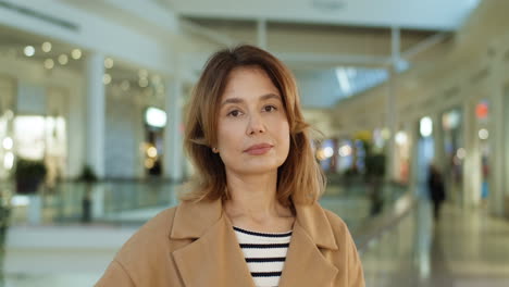 Portrait-Of-Attractive-Woman-In-Coat-Standing-At-Shopping-Mall-Passage,-Turning-Face-And-Smiling-Cheerfully-To-Camera