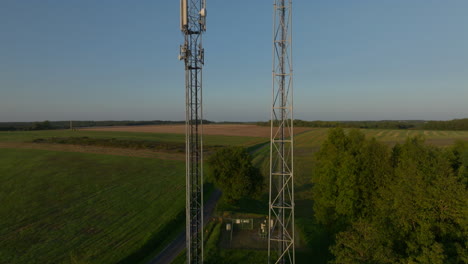 Two-radio-towers-in-the-middle-of-farmland-during-sunrise,-aerial-orbital-dolly-in-tilting-upward