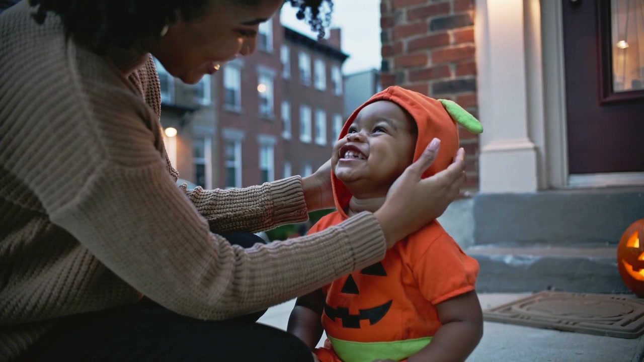 Mamma in maglione che si allunga per mettere un cappuccio a forma di zucca su un bambino piccolo sul portico per Halloween, con zucca illuminata