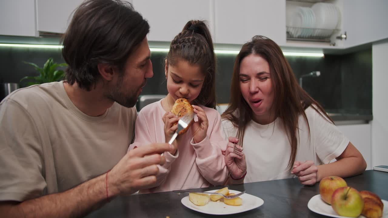 una chica morena con ropa rosa junto con su madre morena en una camiseta blanca y un padre moreno con paja en una camisa beige y desayunando por la mañana en la mesa de comedor en un apartamento moderno en la cocina