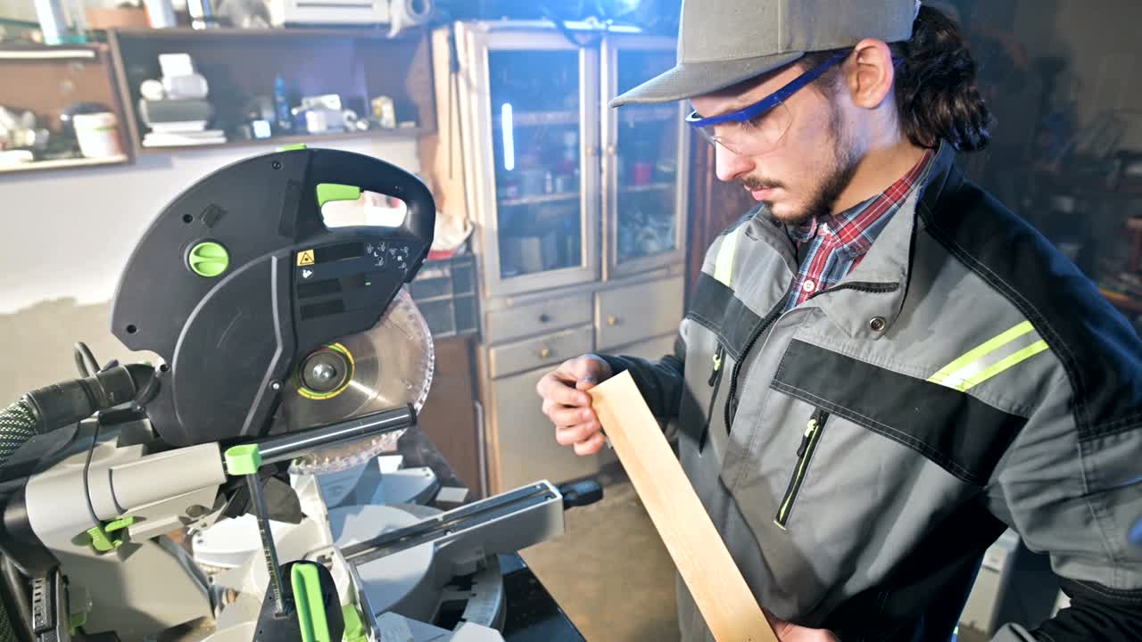 A young man with a beard in gray overalls by profession a carpenter works with a circular cutting machine in his home workshop