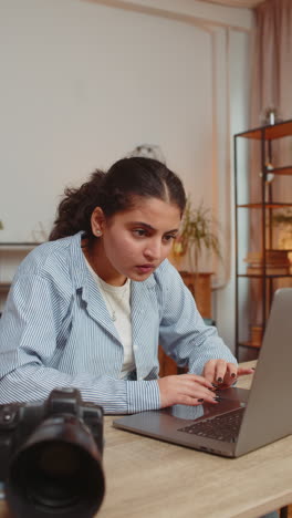 young indian woman photographer transfer data files from camera to laptop at home office table