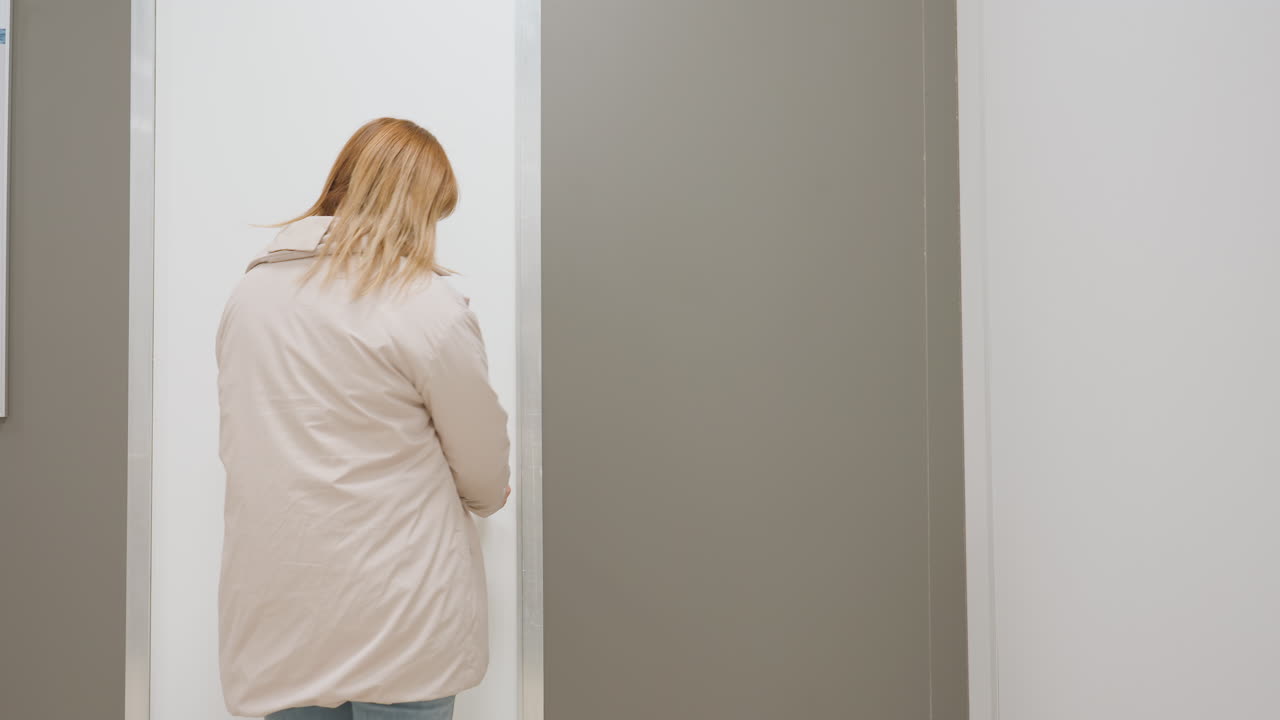 Door opens as elegant woman dressed in jeans, yellow top, and jacket steps out calmly into corridor, holding door handle, indoor office setting with light switch visible on side wall