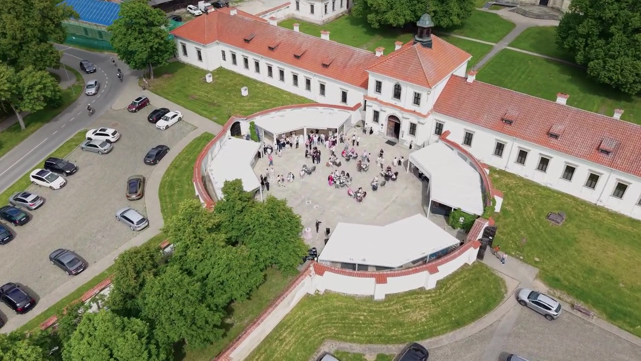 Un video di drone riprende il Monastero di Pazaislis, una vegetazione lussureggiante, un parcheggio pieno e una folla vivace nel cortile d'ingresso in una giornata luminosa. I dettagli architettonici e paesaggistici sono chiaramente visibili