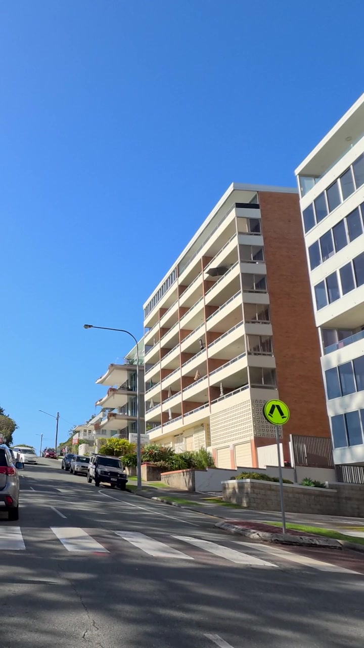 Toma de seguimiento en ángulo bajo de apartamentos modernos, pinos de la Isla Norfolk y coches bajo cielos azules.
