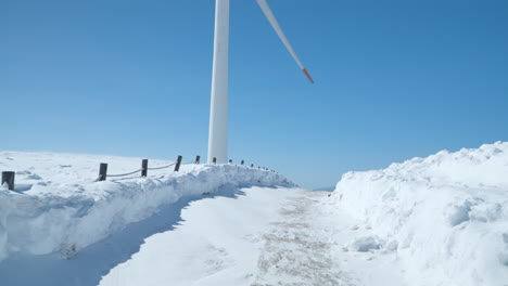 Punto-De-Vista-Del-Conductor-Conduciendo-Por-Una-Carretera-De-Montaña-Cubierta-De-Nieve-Con-Un-Día-Soleado-Con-Cielo-Despejado,-Una-Enorme-Turbina-Eólica-Con-Palas-Giratorias