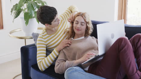 Happy-caucasian-lesbian-couple-lying-on-sofa,-using-laptop-and-smiling-in-sunny-house