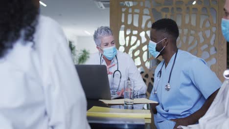 caucasian female senior doctor and diverse hospital colleagues wearing face masks talking at meeting