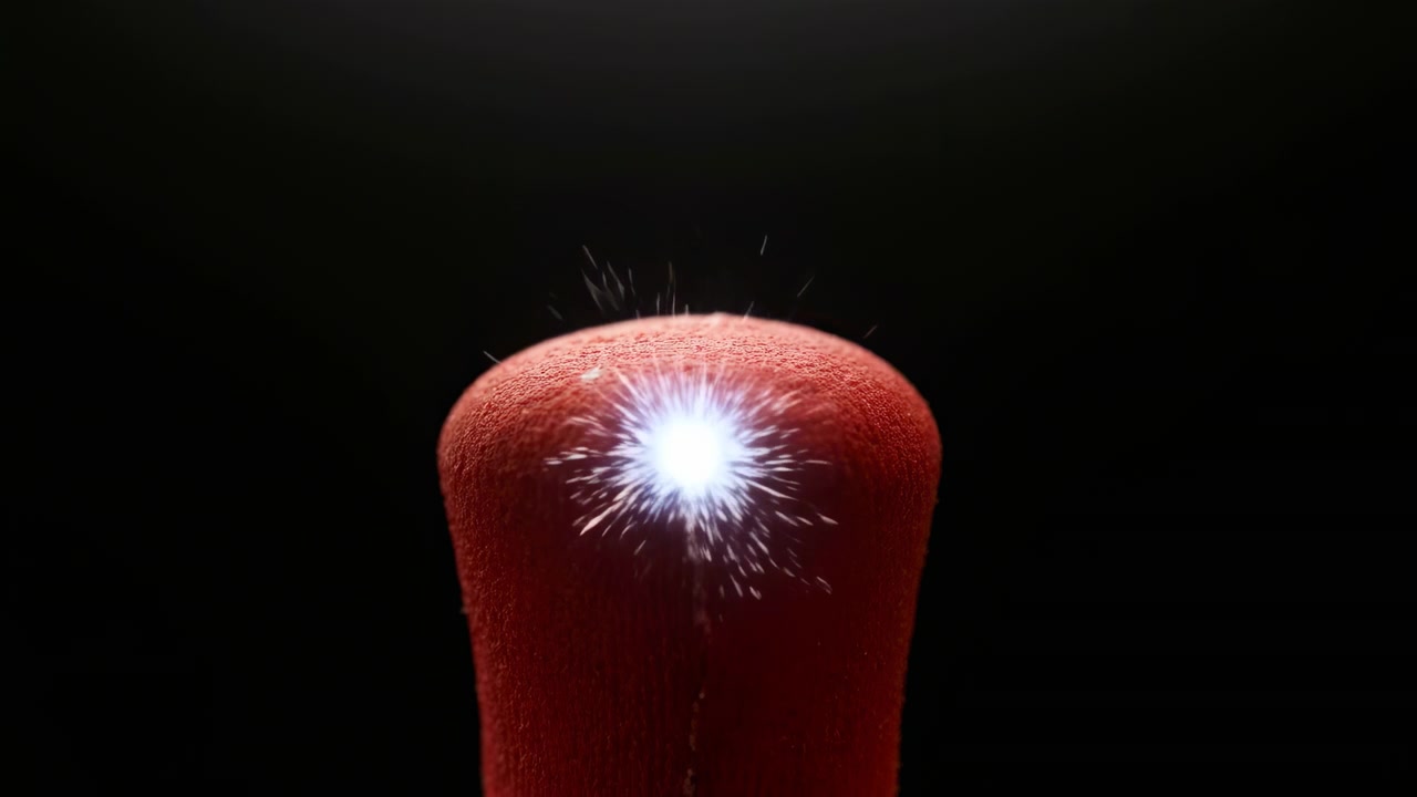 Bright spark appears on textured red match head, turning into glowing orange ignition and rising flame that wraps around rounded tip against a dark, uncluttered background.