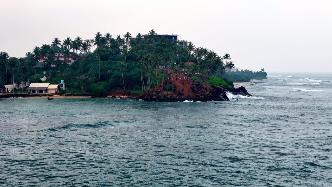 Aerial Telephoto shot from the choppy Indian Ocean captures Parrot Rock, Mirissa, Sri Lanka under a grey overcast sky. Dark palms crown the headland above grey-blue waves