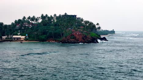 aerial telephoto shot from the choppy indian ocean captures parrot rock, mirissa, sri lanka under a grey overcast sky. dark palms crown the headland above grey-blue waves