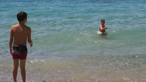 Medium-100fps,-slow-motion-footage-of-caucasian-boy-throwing-pebbles-towards-his-brother,-playing-at-the-seashore,-enjoying-summer-vacations