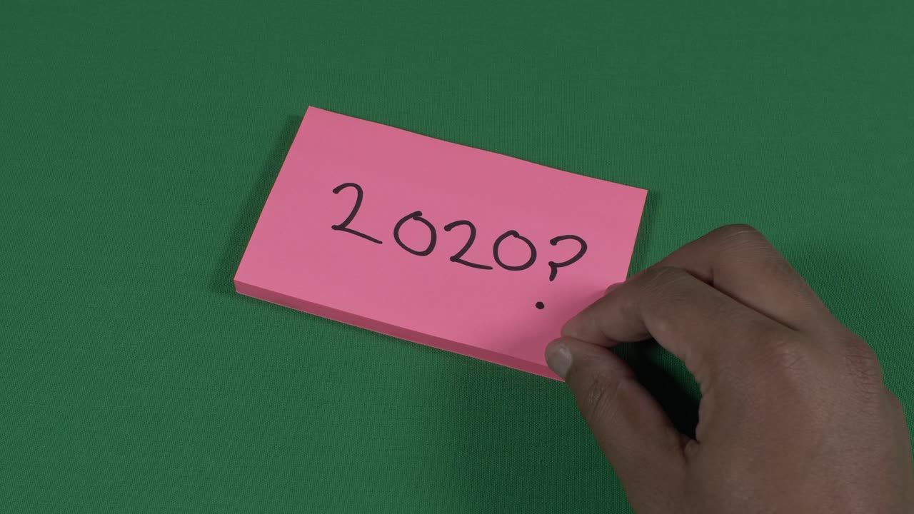 Mano sinistra maschile che scrive 2020 con un punto interrogativo su un post-it rosa