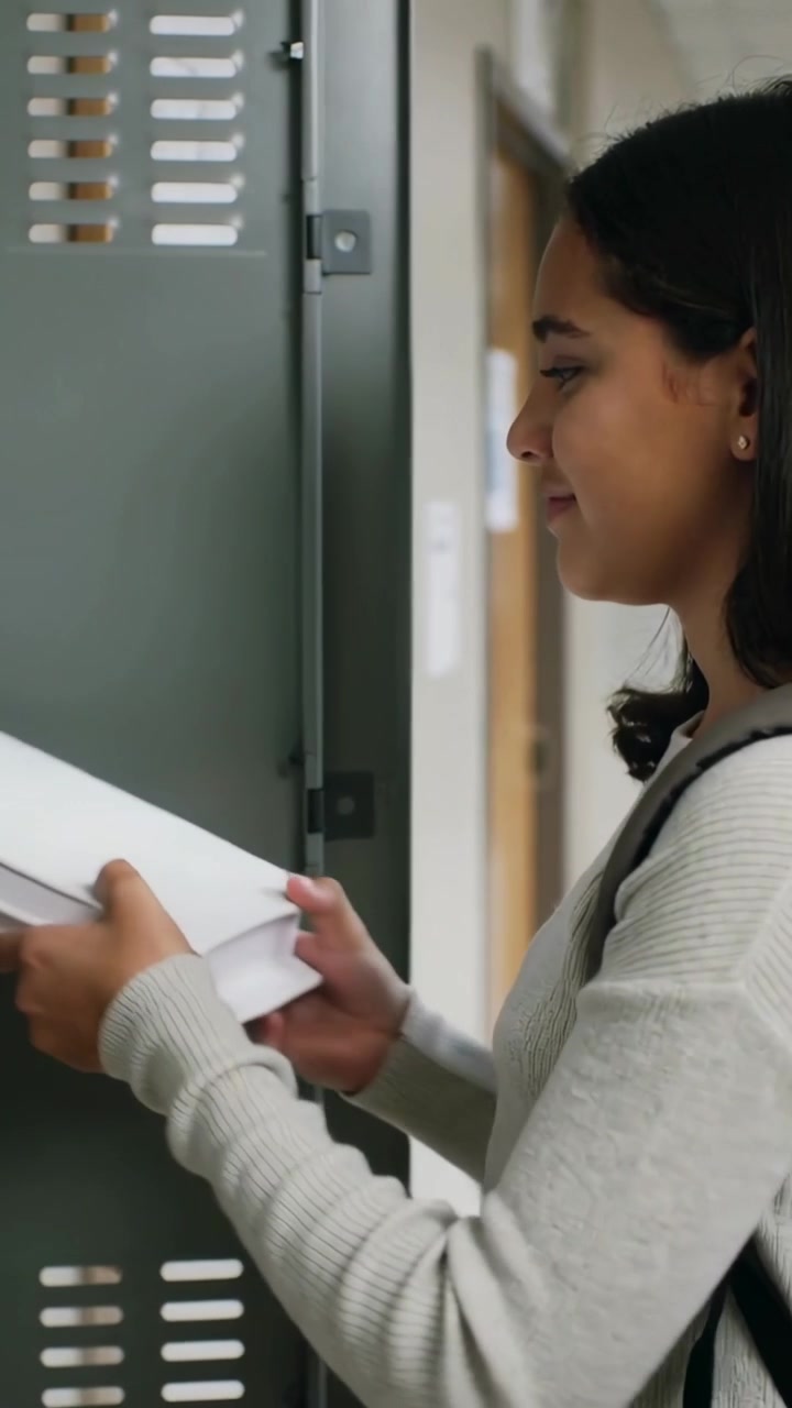 Vídeo vertical: Adolescente de suéter lendo avisos fixados, abrindo armário na escola e organizando livros.