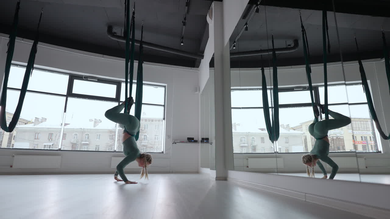 A young beautiful woman performs exercises on a hammock from aero yoga. A woman in the afternoon in front of the mirror in the hall performs stretching alone