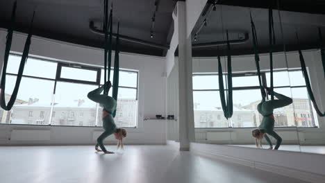 a young beautiful woman performs exercises on a hammock from aero yoga. a woman in the afternoon in front of the mirror in the hall performs stretching alone