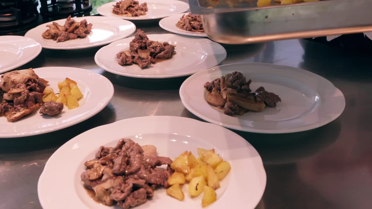 A medium shot captures a chef plating a dish of tender meat with mushrooms and roasted potatoes from a tray onto white plates in a busy kitchen setup.