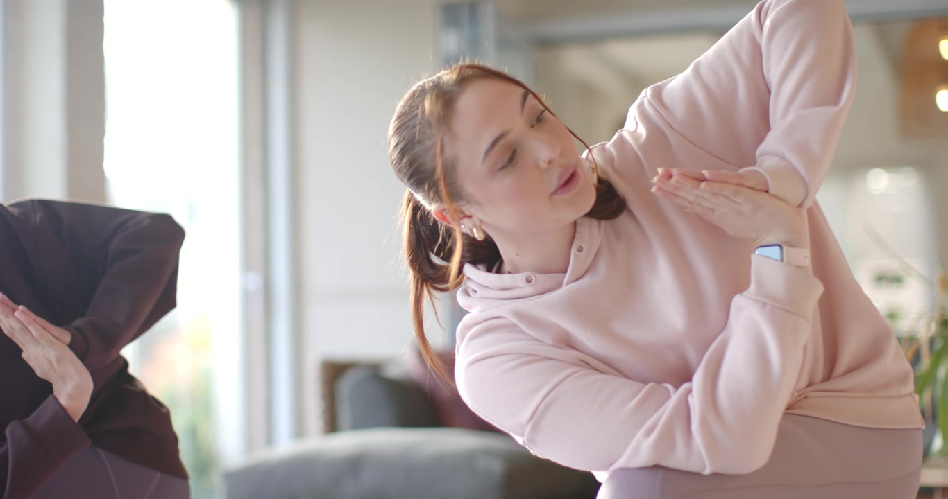 Female friends following cue, doing side-bend while breathing in living room with hoodie