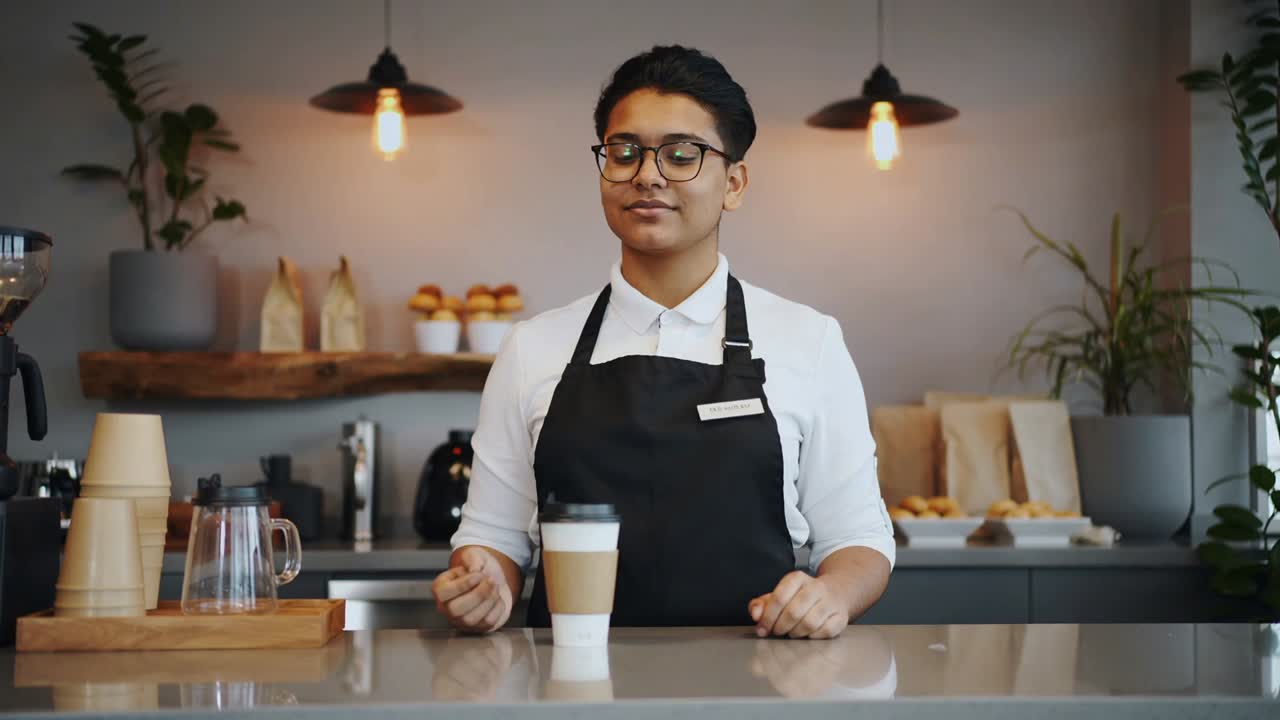 Handing barista wearing white shirt, black apron serving paper cup at counter as customer reaches