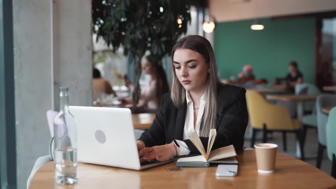 joven, hermosa mujer sentada en un café, vestida con ropa de negocios, se centra atentamente en su portátil con un cuaderno abierto