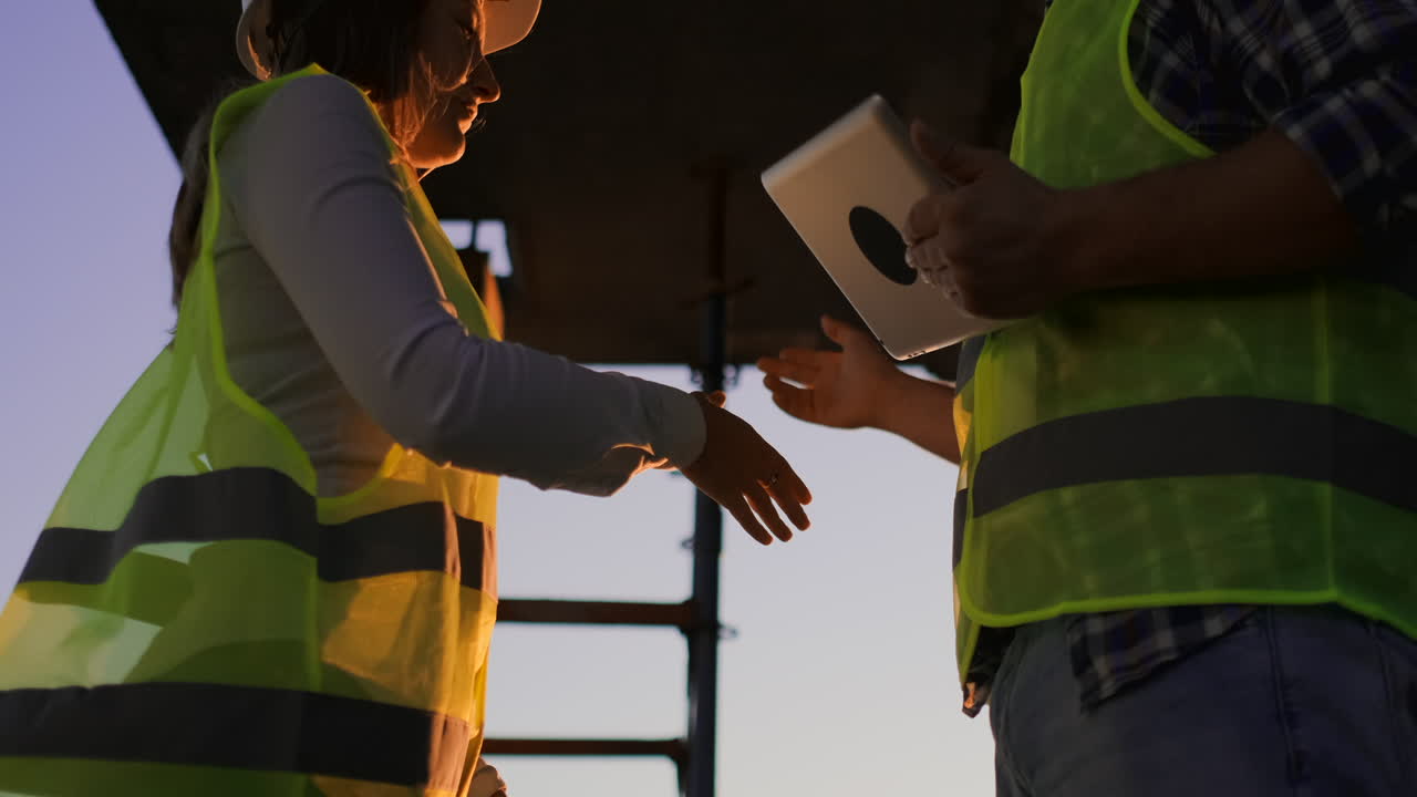 Handsome construction man and woman workers in protective helmets and vests are shaking hands while working in the office center