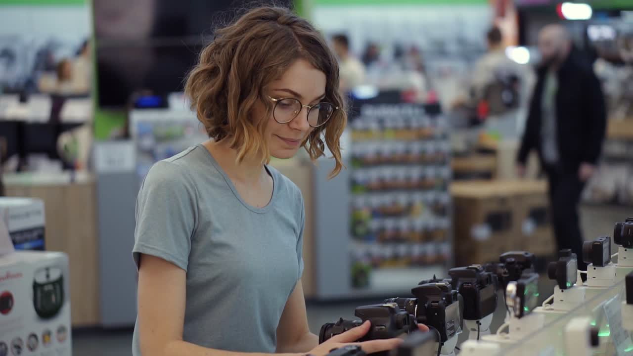 Young woman in glasses shopping for a new photocamera in the electronics store. Trying to decide on the best model. Have Doubts. Checking one camera in hands