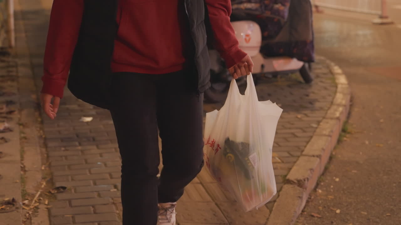 Persona per strada di sera che porta una busta di plastica della spesa, marciapiede urbano con moto parcheggiata e riflessi al neon sulla pavimentazione bagnata, giacca rossa e scarpe da ginnastica visibili in un'inquadratura ravvicinata della parte inferiore del corpo, solitaria.