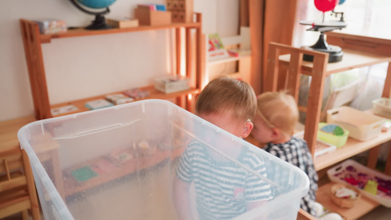 Junge im gestreiften Hemd beobachtet aufmerksam das Innere eines transparenten Gummi-Behälters, während ein kleines Mädchen in einem hellen Klassenzimmer voller Regale, Bücher und einem Globus weggeht
