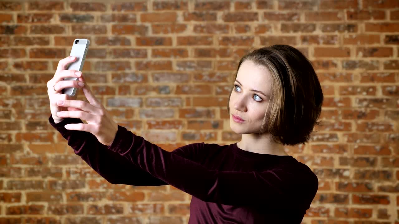 joven hermosa está haciendo selfie en su teléfono inteligente, sonriendo, concepto de comunicación, fondo de ladrillo