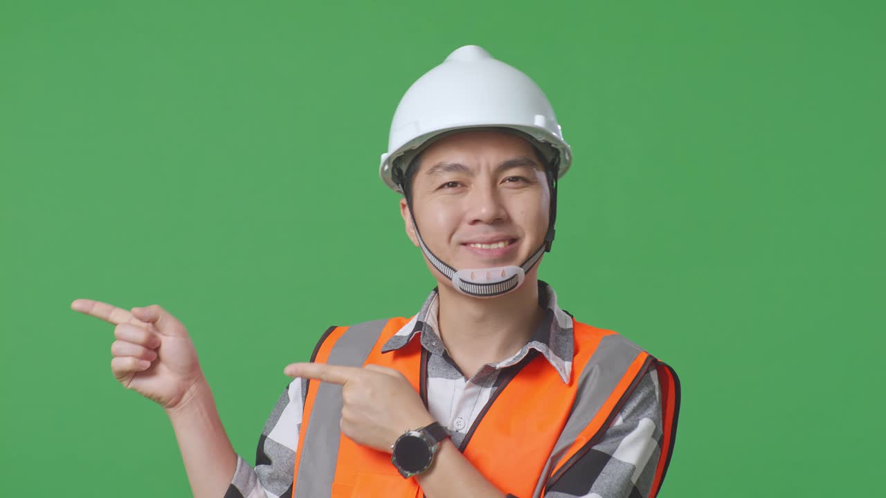 Close Up Of Asian Male Engineer With Safety Helmet Smiling And Pointing To Side While Standing In The Green Screen Background Studio
