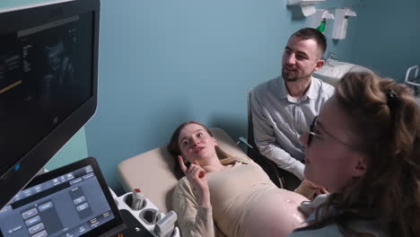 female gynecologist showing image of fetus on screen of ultrasound machine to happy future parents 1