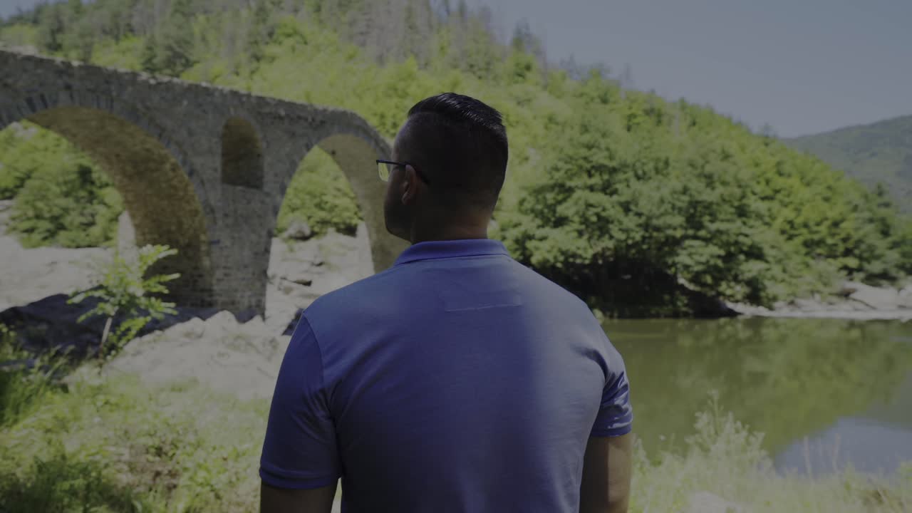 Panning around a tourist who is admiring the architectural design of the Devil's Bridge and beautiful scenery of Arda River right next to the Rhodope Mountains in Bulgaria