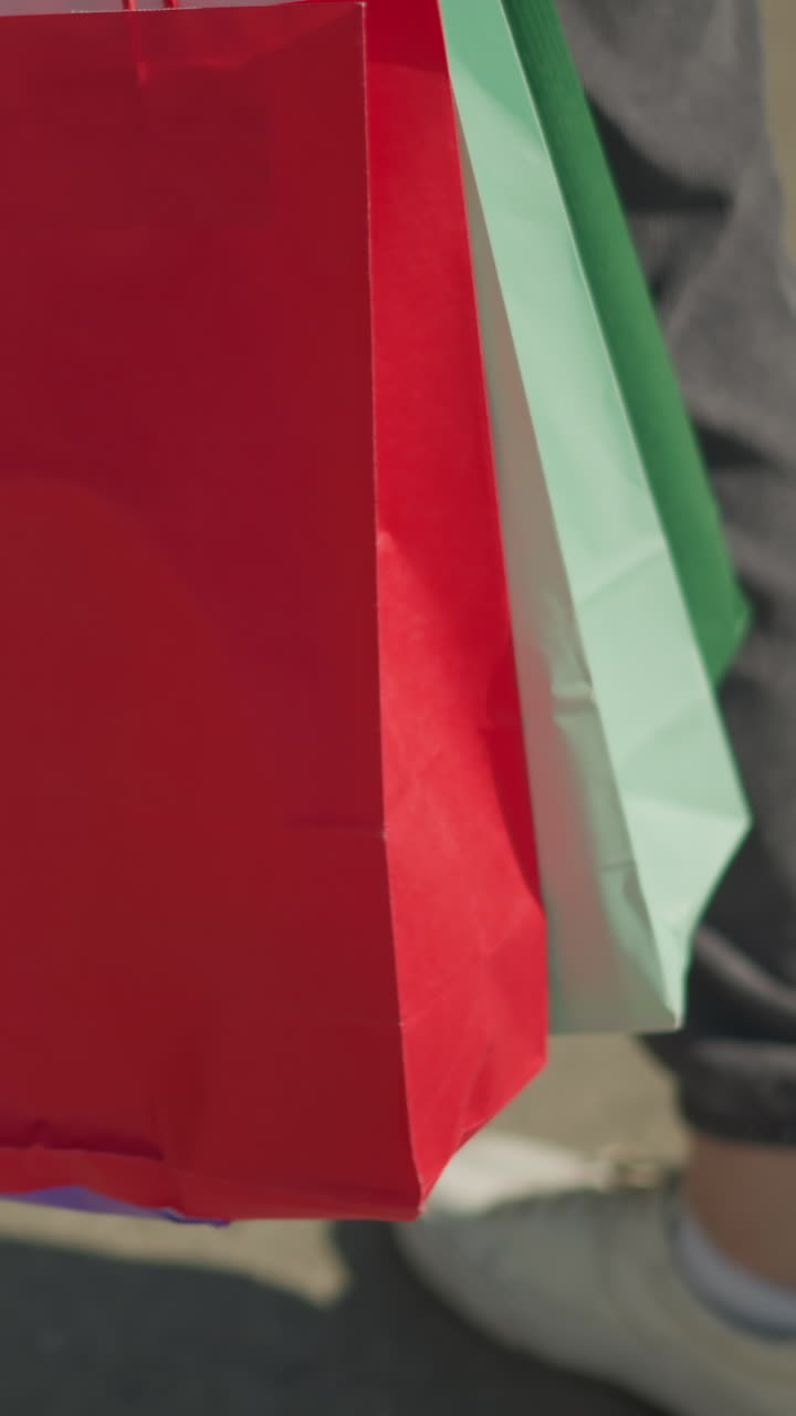 vista de la pierna de individuos que usan corredores grises y zapatos de lona blancos caminando sobre alquitrán, llevando bolsas de compras verdes, menta y rojas con una sombra visible en el suelo