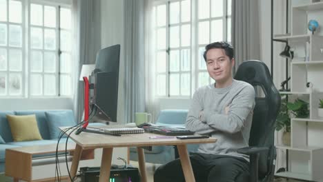 asian man in long sleeved t-shirt and black pants crossing his arms and smiling to camera while working with desktop computer at home.