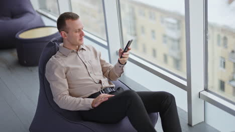 calm relaxed man sits in bean bag chair, speaks and types something on his phone. view from above at the backdrop of panoramic window.