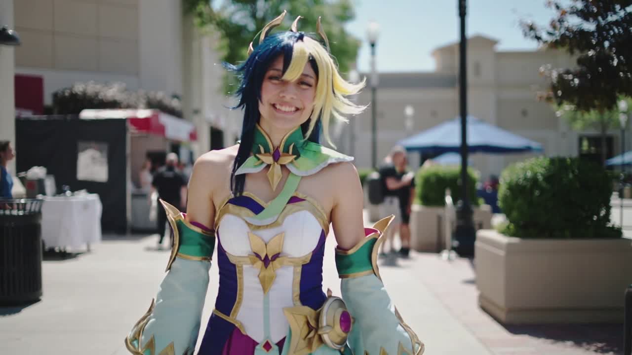 Cosplayer caminhando na praça, posando com armadura, peruca e manoplas, enquanto a câmera ajusta o enquadramento.