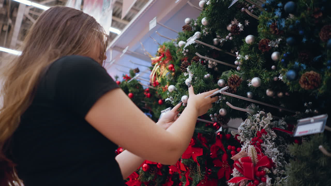 primer plano de una mujer joven de cabello largo y marrón con camisa negra y gafas de sol admirando las coronas de navidad decoradas con adornos de plata, piñas y cintas festivas en el pasillo de la tienda de vacaciones