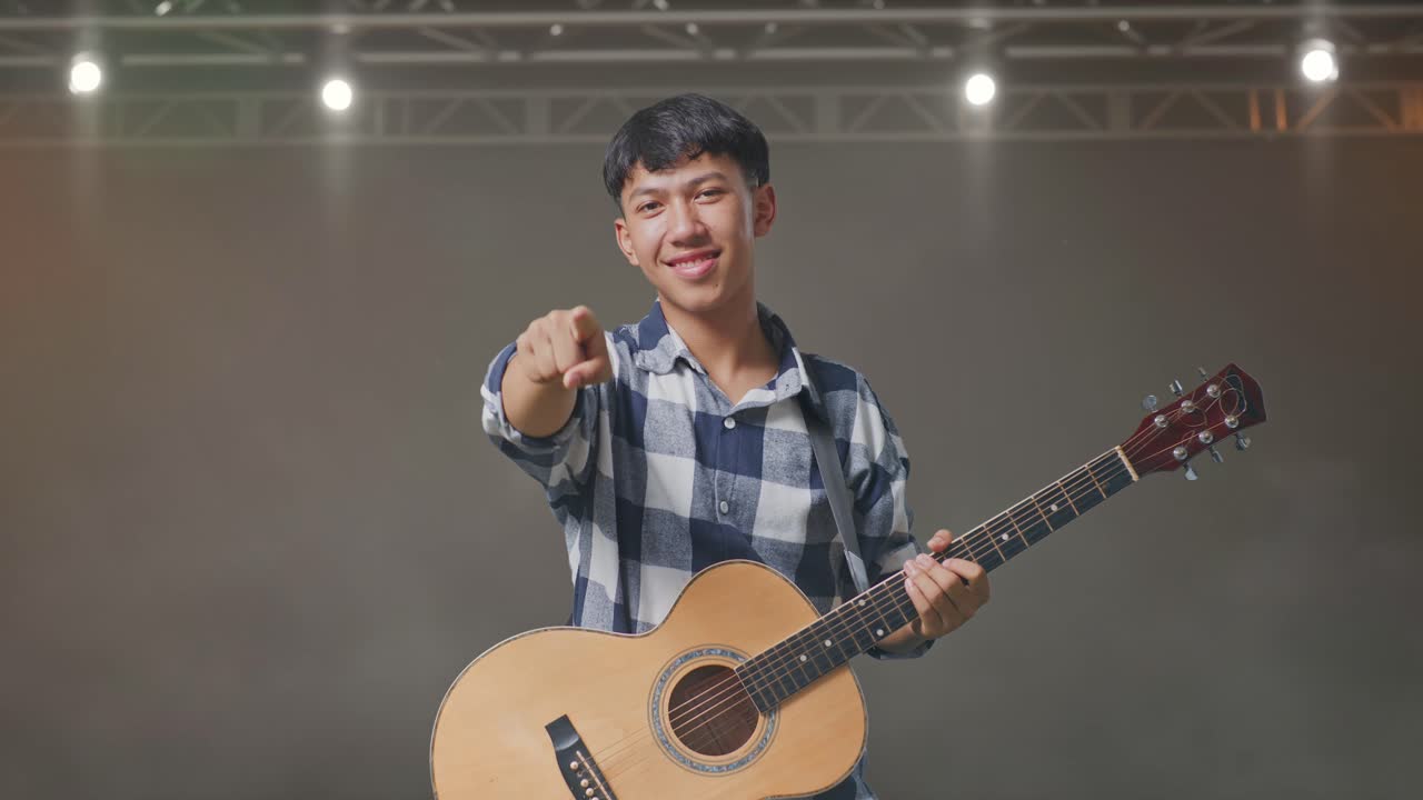 기타를 들고 있는 아시아인 십대가 무대에서 정직한 몸짓을 하고 미소 짓고 있습니다.