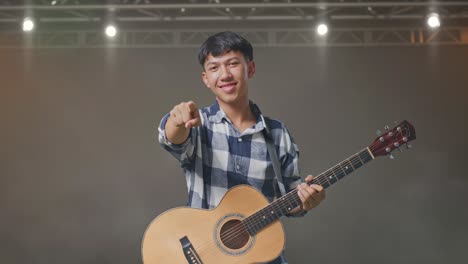 adolescente asiático con guitarra haciendo un gesto honesto y sonriendo en el escenario